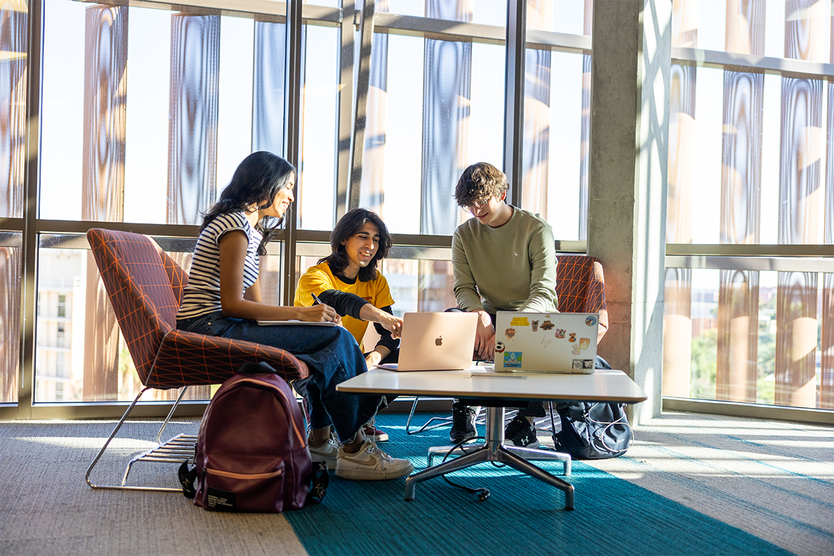 Three students study together at a table with laptops in a sunlit room.