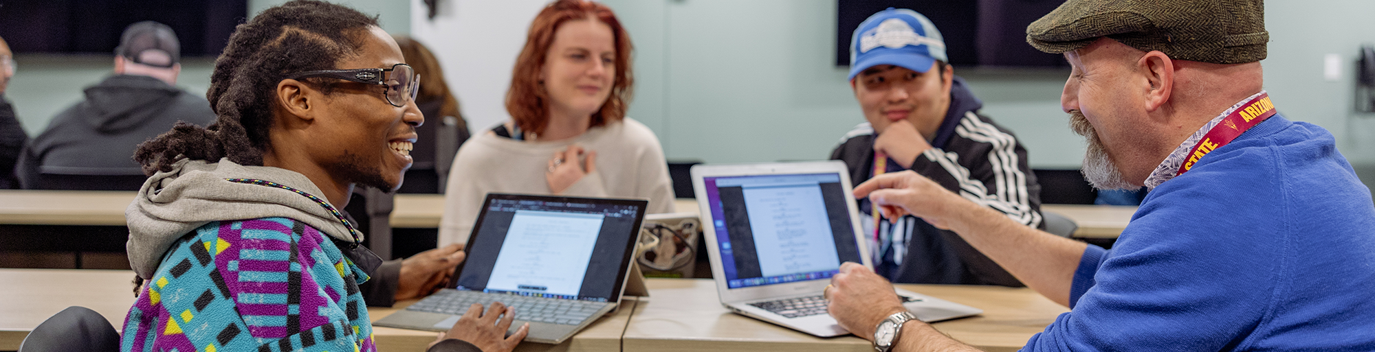 Group of people having a discussion with open laptops in a classroom setting.