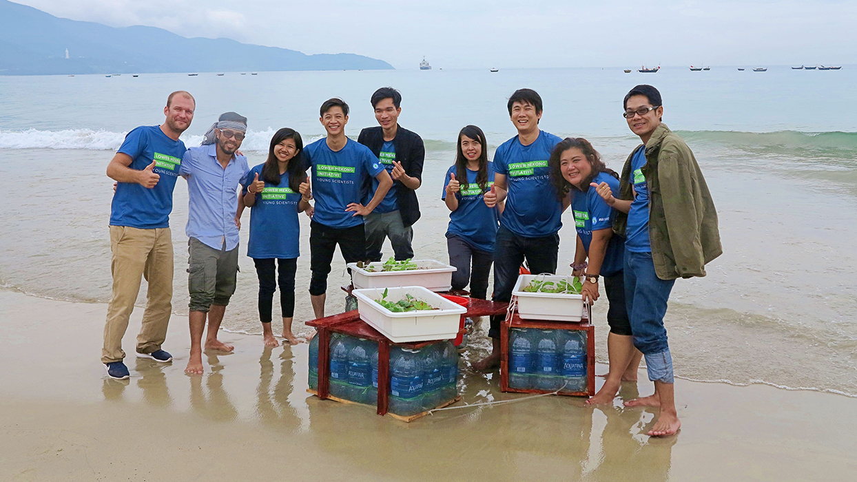 A group of eight people on a beach with plant trays and water containers, smiling and giving thumbs-up.