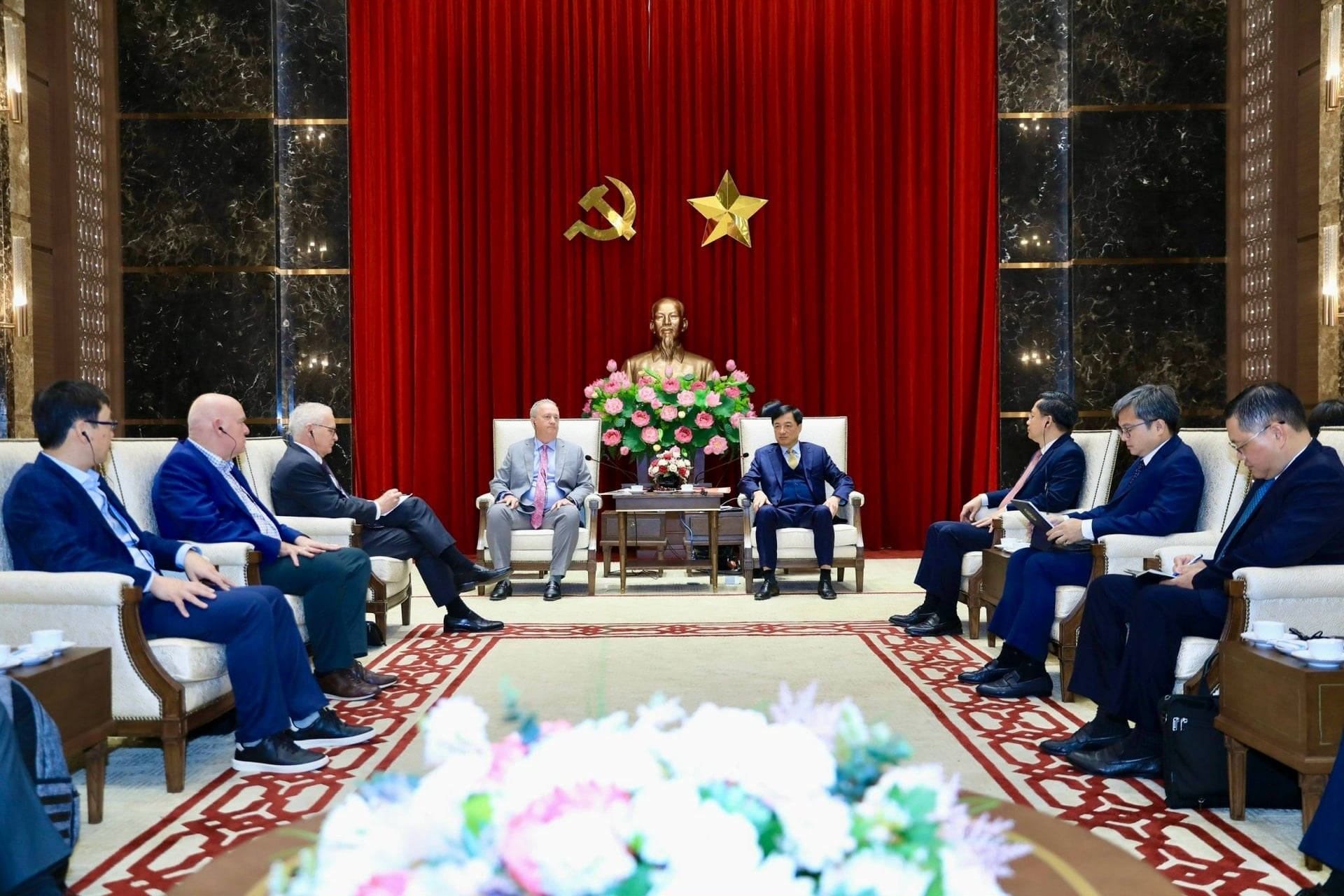 A formal meeting room with red curtains, a central bust, and several people sitting in two rows facing each other.