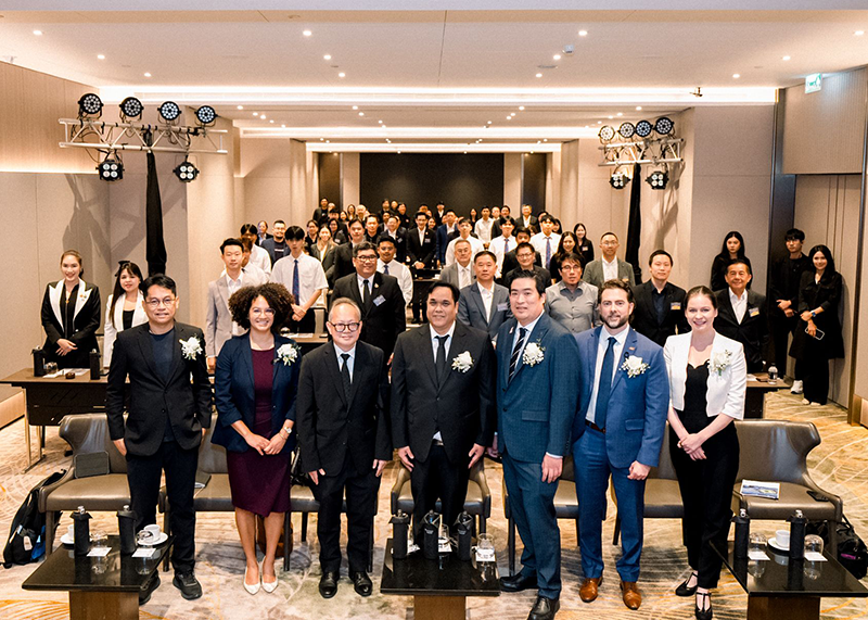 A formal group photo in a modern auditorium with seven people standing in the foreground and a seated audience behind them.