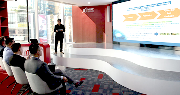 A speaker presents in a modern conference room with an audience and a screen detailing the Semiconductor Implementation Academy.