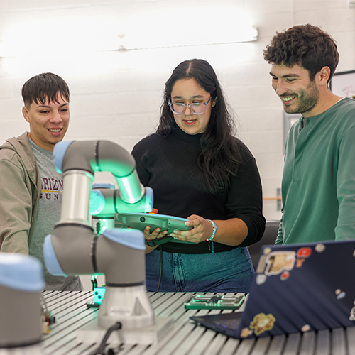 Three young adults, one female and two males, collaborate over a tablet in a tech workspace, with a robotic arm nearby.