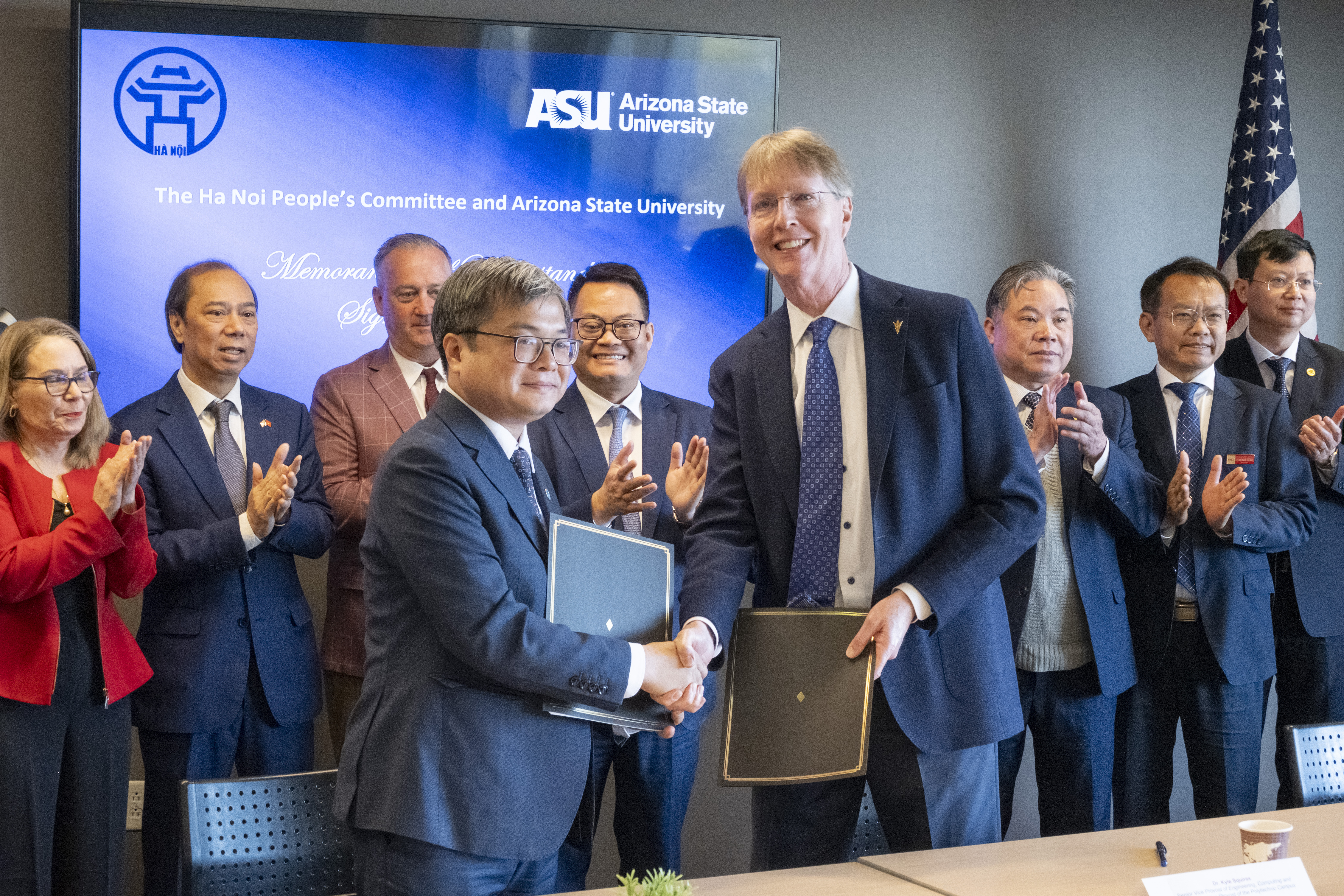 Two men in suits shake hands holding documents, in front of a digital screen and clapping attendees.