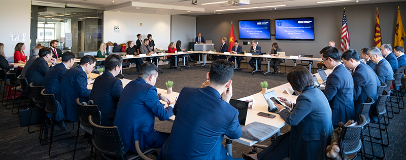 A business meeting in a modern conference room with a U-shaped table and multiple attendees around it.