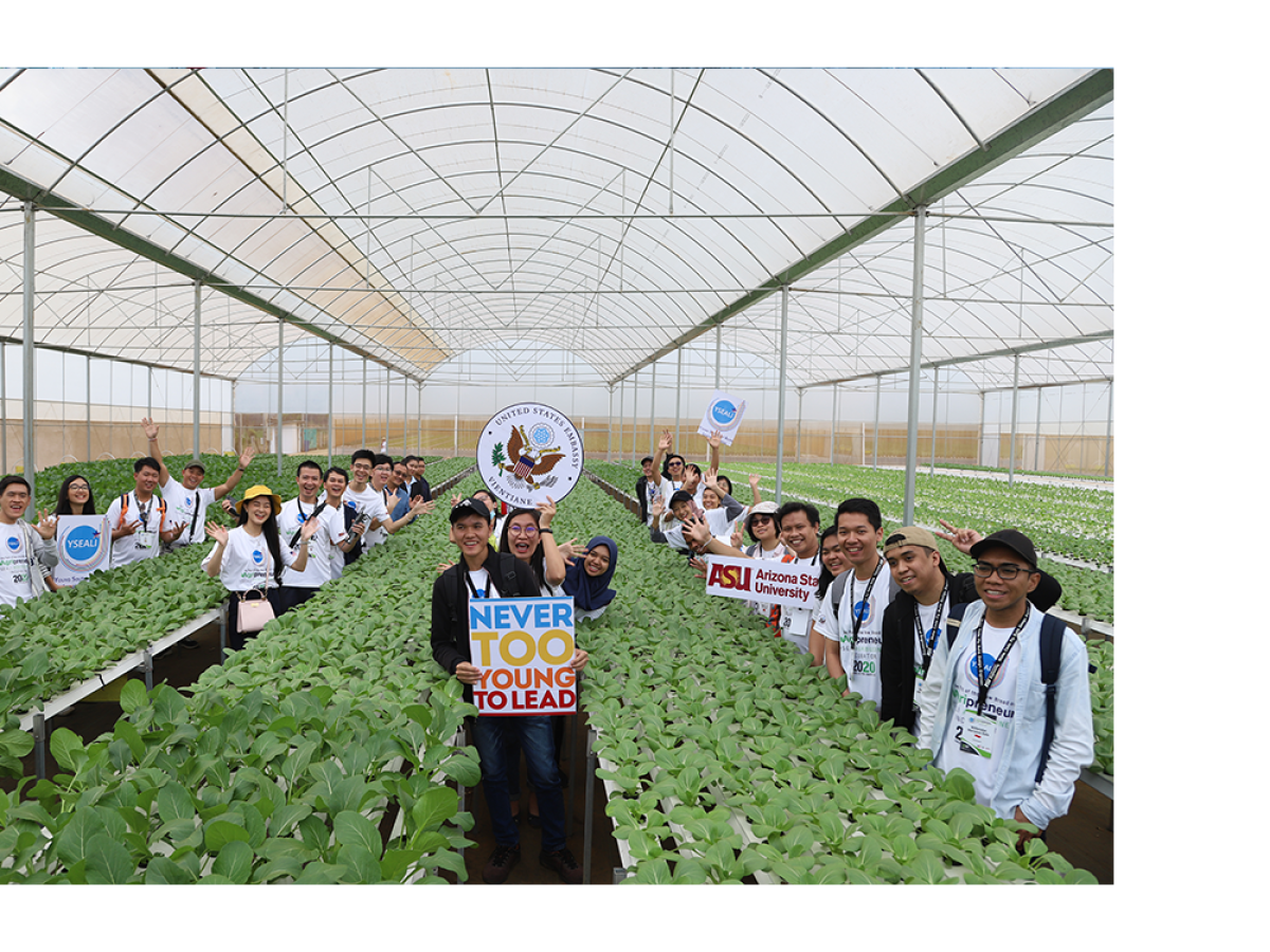 A group of people in a greenhouse, holding signs among rows of green plants.