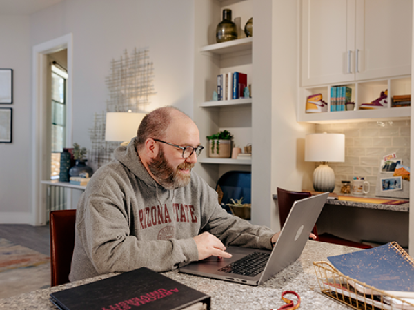 A man in a gray hoodie working on a laptop at a kitchen island, surrounded by a well-decorated workspace with bookshelves and a lamp.