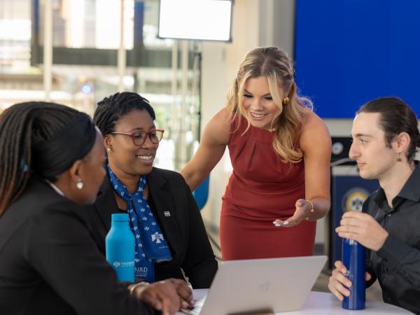 A group of four people collaborates around a laptop in a bright, modern workspace. A woman in a red dress gestures animatedly while speaking to her colleagues.